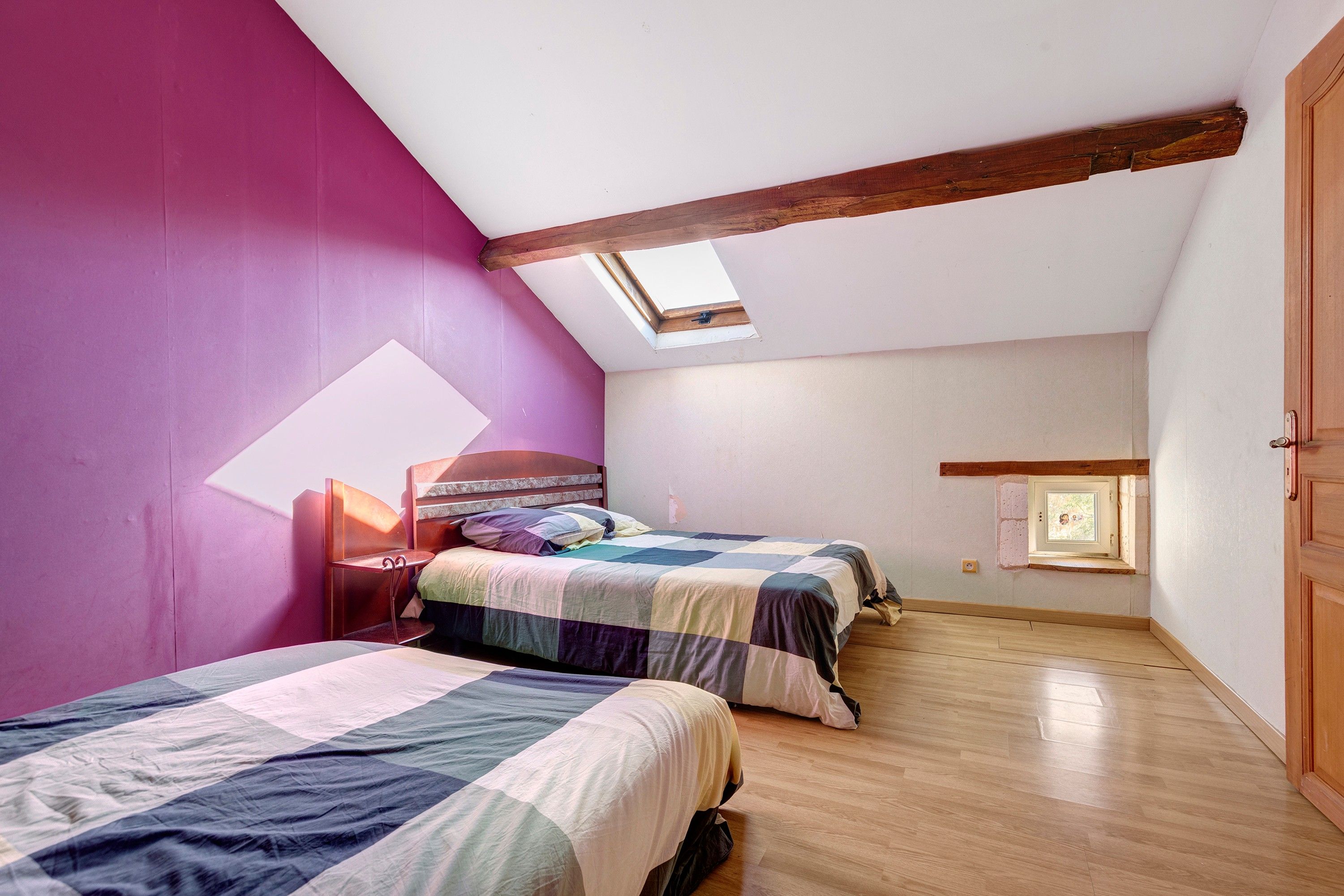 A bedroom with two beds, a skylight, wooden beams, and a small window.