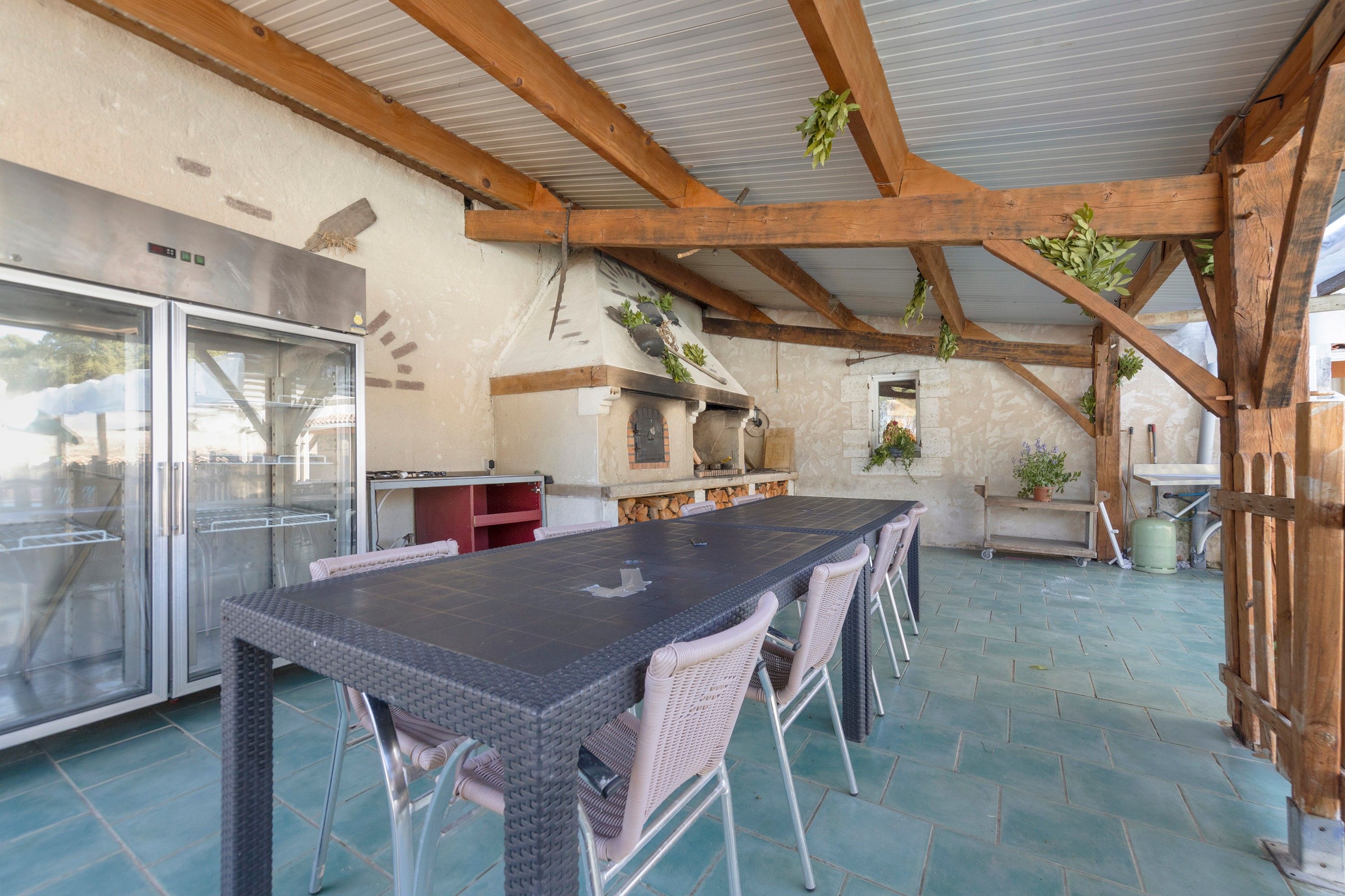 An outdoor kitchen area with a long table and chairs, a refrigerator, and a stone fireplace.