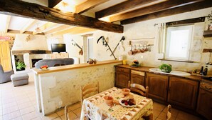 A rustic kitchen with wooden beams, a dining table set with bowls and fruits, and a fireplace in the background.