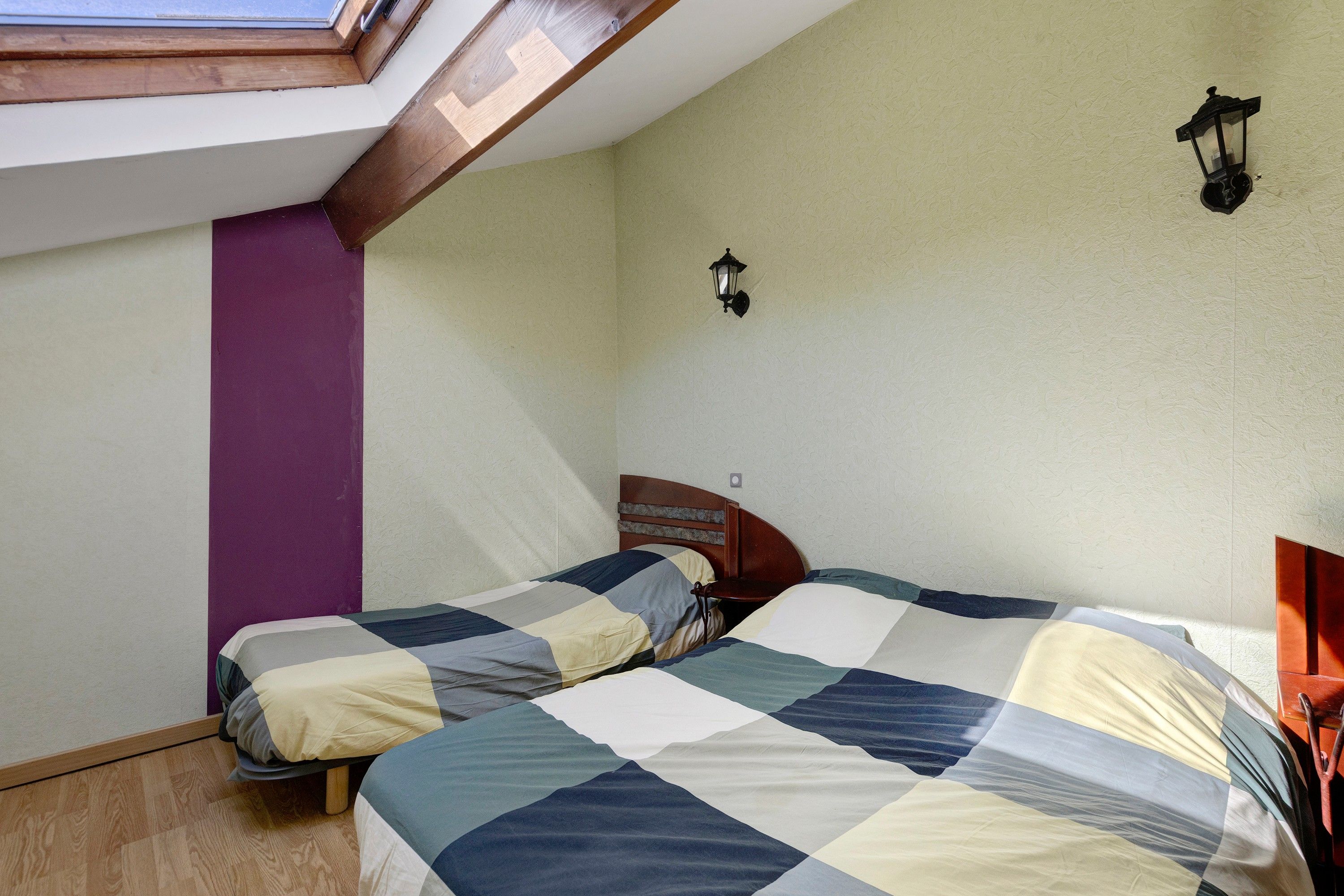 A room with two beds, a skylight, a wall-mounted light fixture, and a wooden headboard.