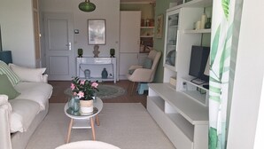 Een woonkamer met een bank, een bijzettafel met bloemen, een televisie en een boekenkast.
