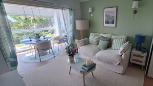 Een woonkamer met een bank, een salontafel, een bijzettafeltje, een lamp, een lijst met een foto en een raam met gordijnen.