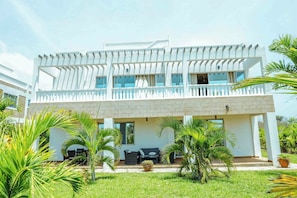 A two-story building with a balcony, a covered outdoor seating area, and a well-maintained garden with palm trees. - Luxurious 5-bedroom Villa With Private Pool in Diani, Kenya (Diani Beach)
