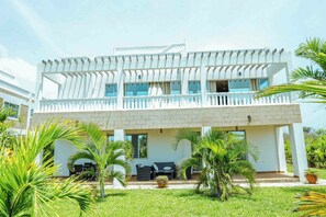 A two-story building with a balcony, a covered outdoor seating area, and a well-maintained garden with palm trees. - Luxurious 5-bedroom Villa With Private Pool in Diani, Kenya (Diani Beach)