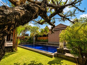 A swimming pool surrounded by lush greenery, a stone pathway, and a traditional building with a tiled roof.