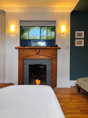 Una habitación con una televisión sobre una chimenea con repisa, una cama con cubrecama blanco y dos lámparas de pared.