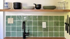 Cozinha com pia preta, backsplash de azulejos verdes e prateleiras de madeira com diversos utensílios de cozinha.