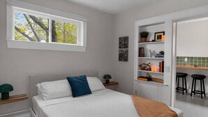 Quarto com cama, mesa de cabeceira, janela, estante com livros e objetos decorativos, e um espaço de cozinha com banquetas.