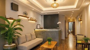 A modern hotel lobby with a sofa, a wooden coffee table, a reception desk, and a hallway leading to other rooms. - Ben Thanh Hideaway - A Cozi Stay  (Ho Chi Minh City)