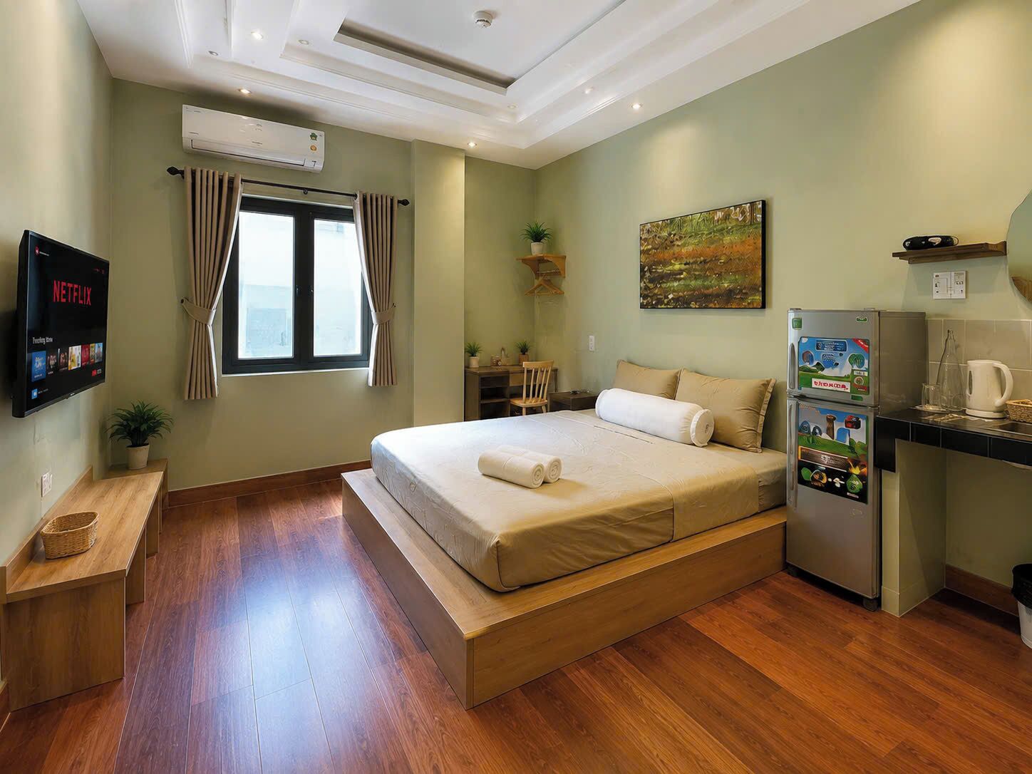 A bedroom with a bed, a TV, a desk, a refrigerator, and a window.