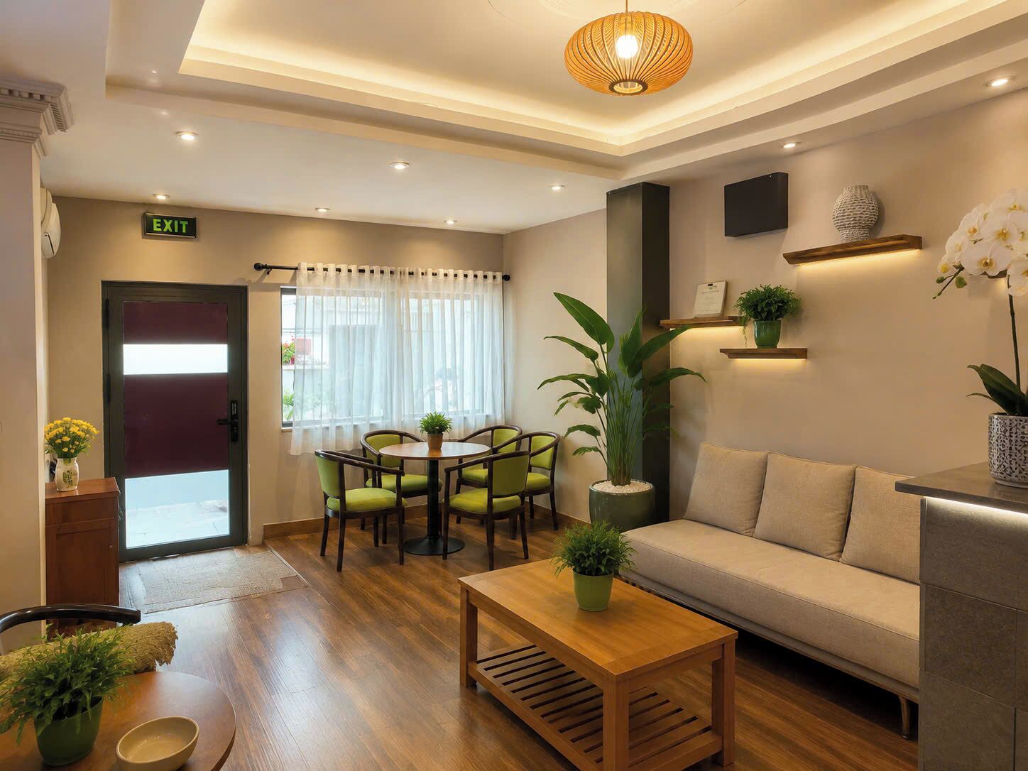 A hotel lobby with a reception desk, a sofa, a dining area with chairs, a large potted plant, and an exit sign.