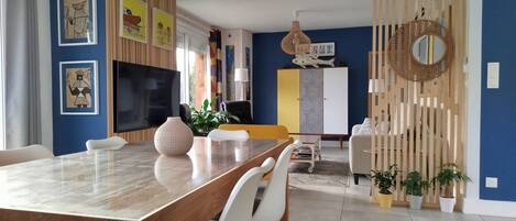A modern living room with a wooden dining table, white chairs, a TV, and a large window.