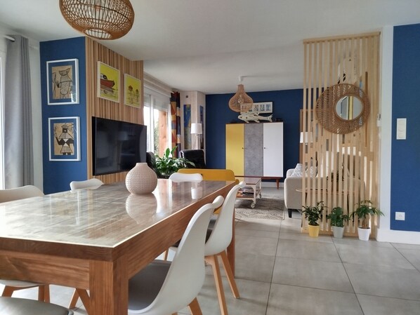 Ein modernes Wohnzimmer mit einem Holzesstisch, weißen Stühlen, einem Fernseher und einem großen Fenster.