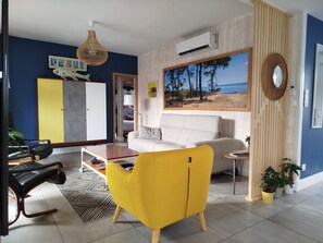 Ein Wohnzimmer mit einer Couch, einem gelben Sessel, einem Couchtisch, einer Pendelleuchte, einer Klimaanlage, einem Strandbild und einem Spiegel an der Wand.