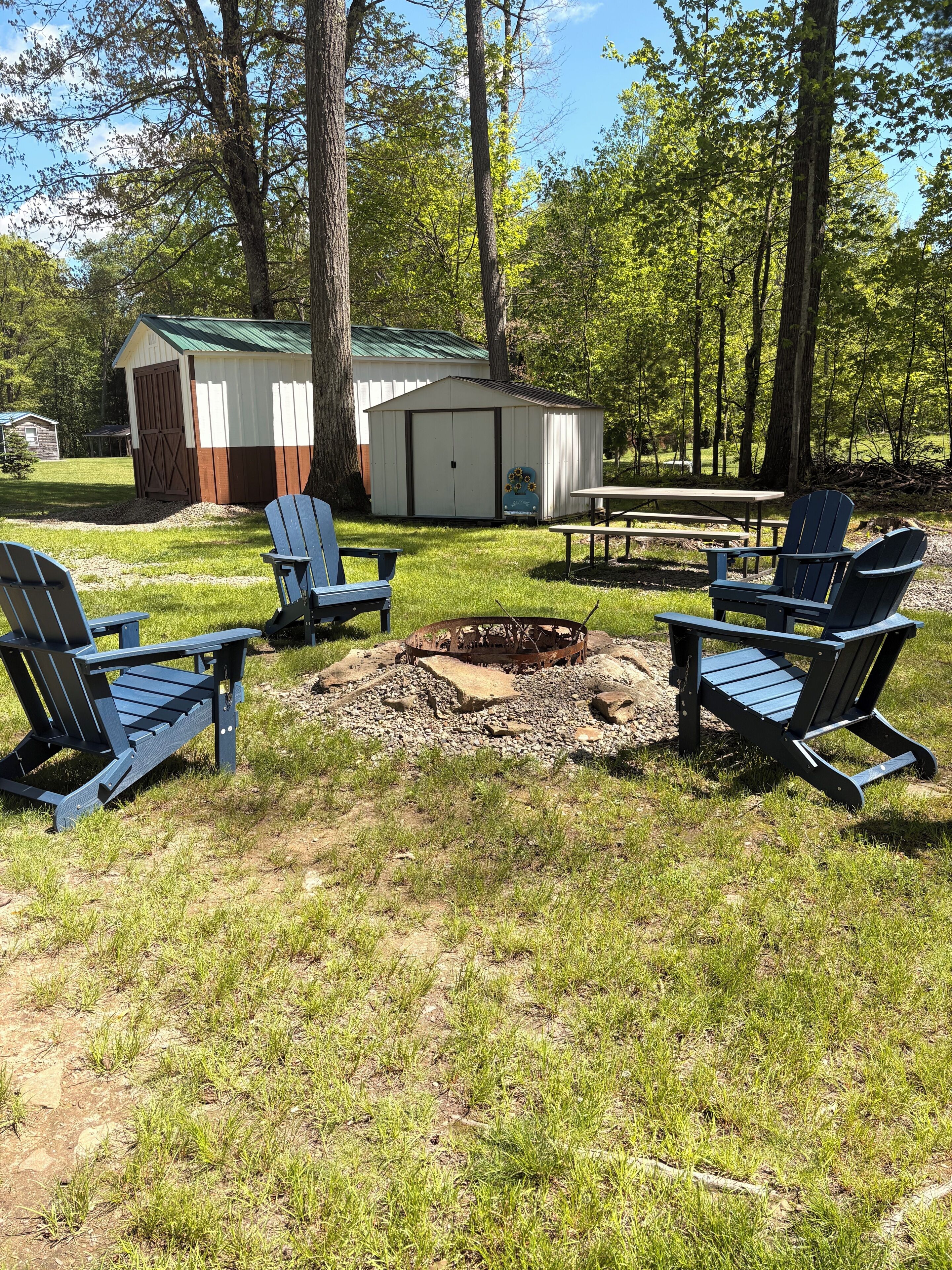 Un terrain de camping équipé de quatre chaises Adirondack, d’un foyer, d’une table de pique-nique et de deux abris de jardin.