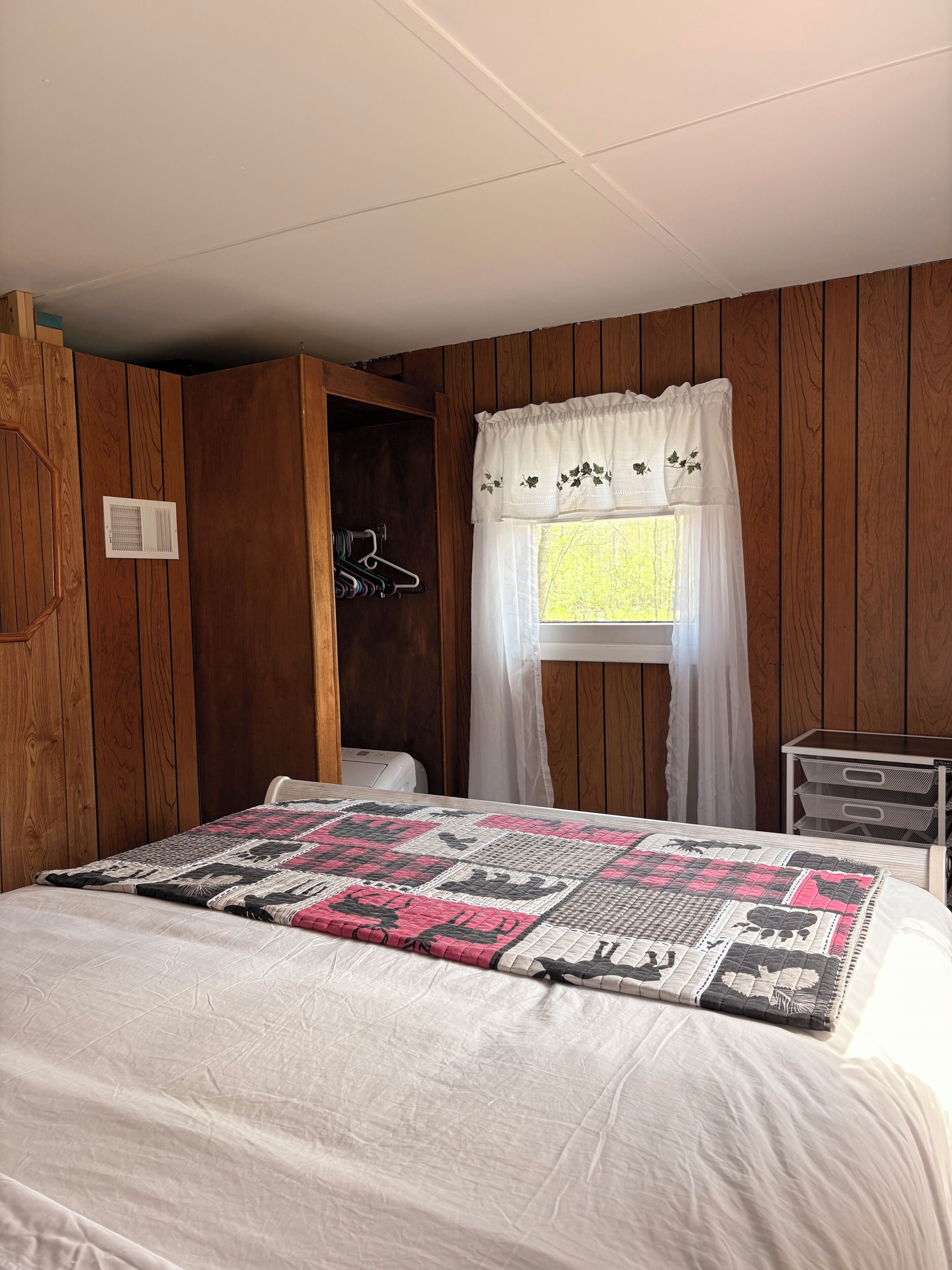Un dormitorio con cama, ventana con cortina blanca, paredes de madera, radiador blanco y una mesita de noche con cajones.