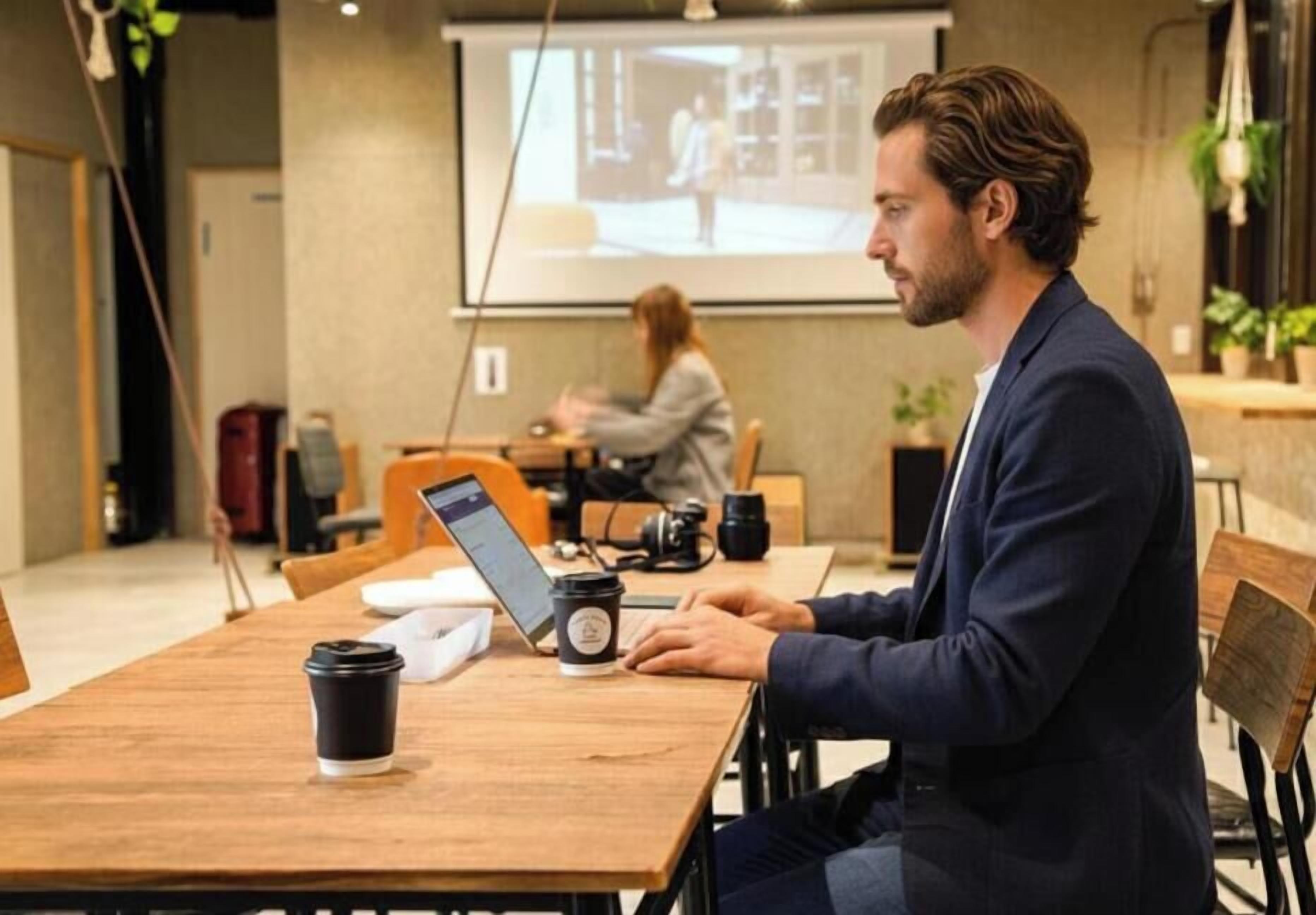 Un hombre sentado a una mesa de madera con una laptop y una taza de café, en una habitación con una pantalla de proyector mostrando una imagen.