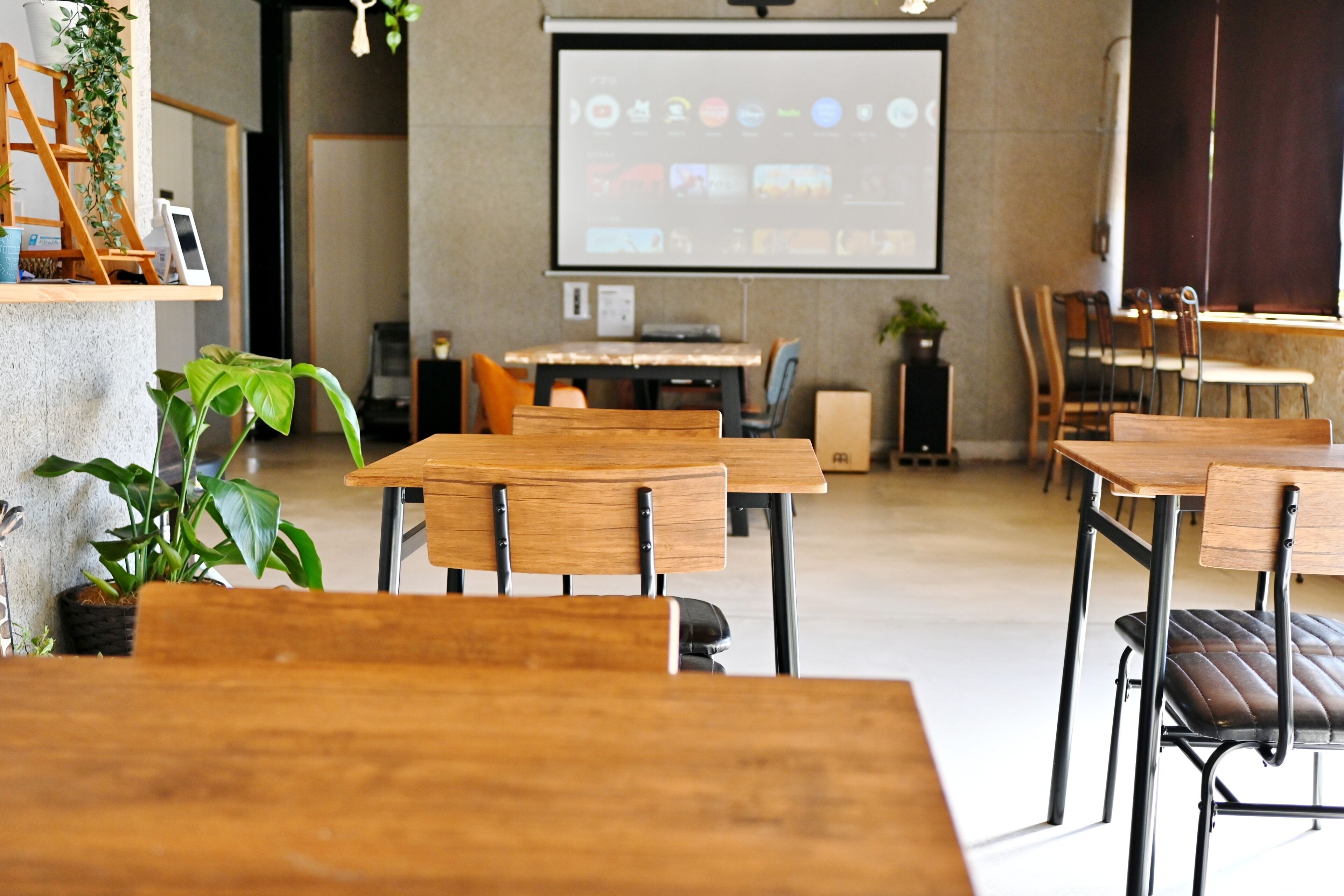 A modern conference room with wooden tables and chairs, a projector screen, and a potted plant.