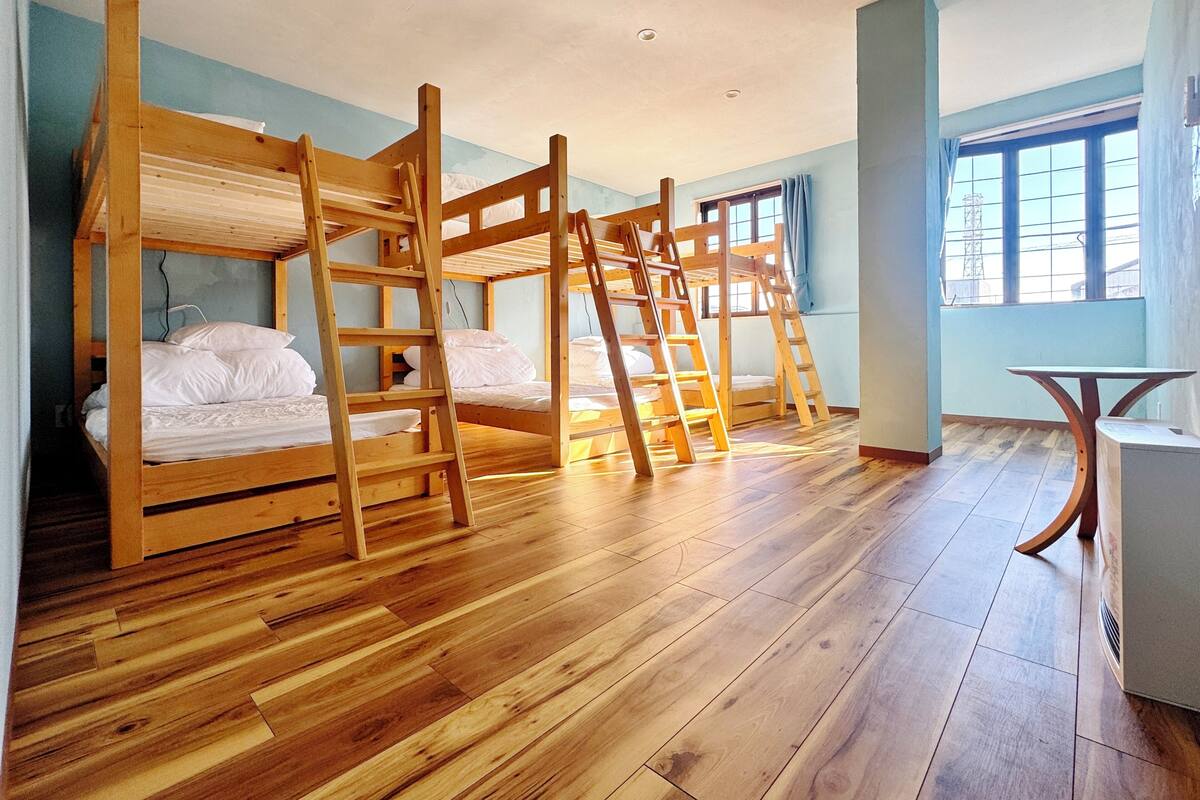 Una habitación con literas, piso de madera, una mesita y una ventana con cortinas.