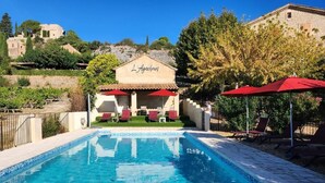 Zona junto a la piscina con un edificio que dice 'L'Agachou', sombrillas rojas y sillones de descanso.