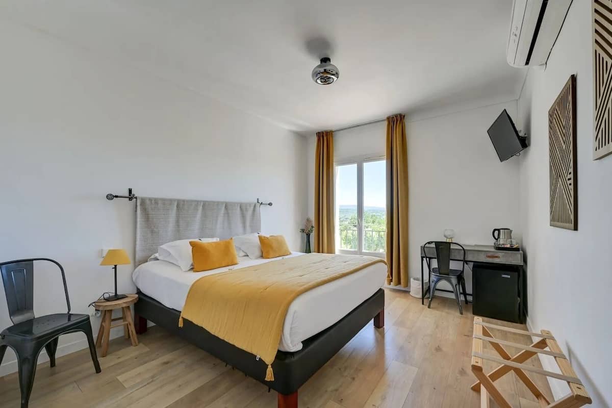 A hotel room with a bed, a desk, a chair, a lamp, and a view of the outdoors.