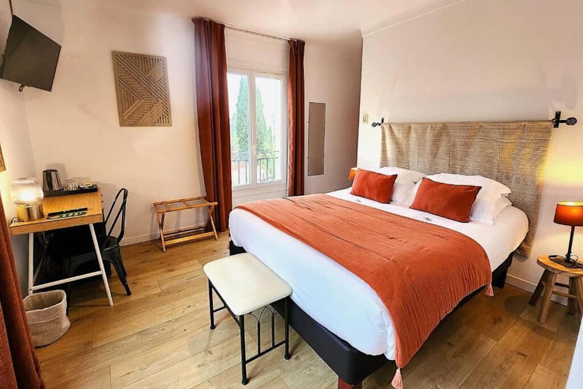 A bedroom with a bed, a desk, a chair, a small stool, a TV, and a window with curtains.