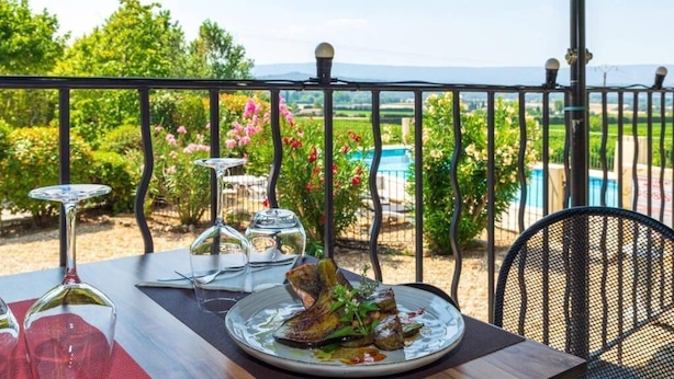 Una mesa de comedor con un plato de comida, copas de vino y una silla, frente a un balcón con vistas a una piscina y vegetación.