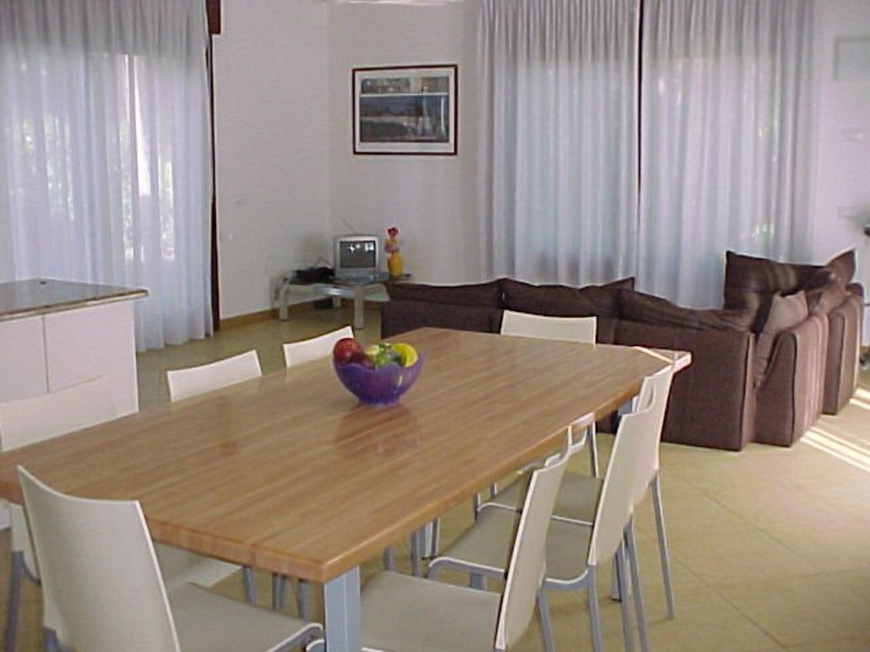 A dining area with a wooden table, white chairs, a brown sofa, and a small television on a stand.