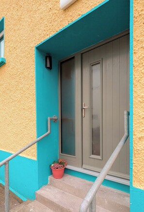 En bygning med gul facade, teal detaljer, en grå dør med glasrude og en lille plantekasse med blomster foran trappen.