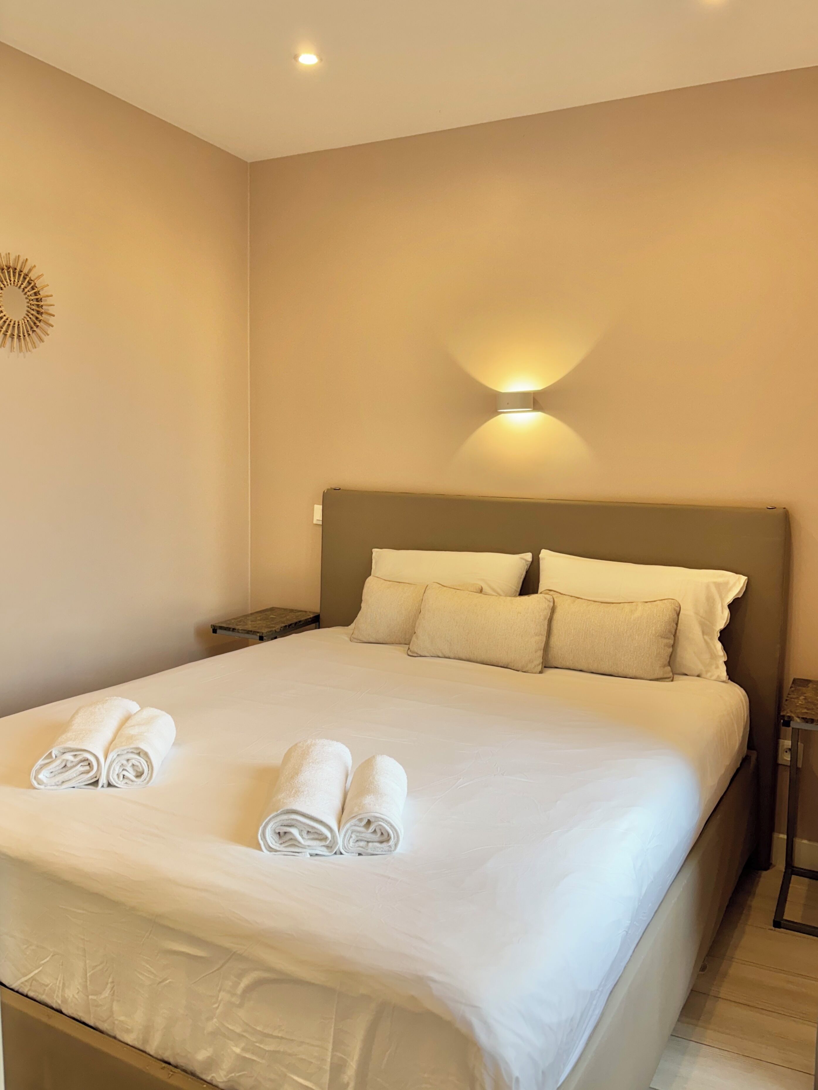 Un letto ben rifatto con cuscini e asciugamani arrotolati, un comodino, una lampada a parete e un elemento decorativo a parete.