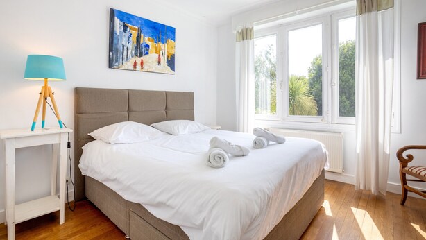Un dormitorio con cama, mesita de noche con lámpara, un cuadro en la pared y ventana con cortinas.