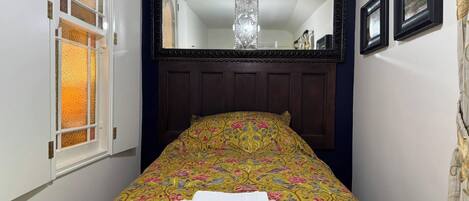 A narrow room with a bed, a large mirror, and a window with stained glass.