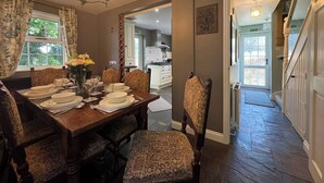 Een eetkamer met een houten tafel voor zes personen, gestoffeerde stoelen, een kroonluchter en uitzicht op de keuken door een open deur.