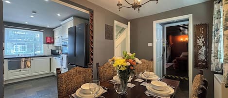 A dining area with a dark wooden table set for six, a chandelier, and a view into the kitchen.