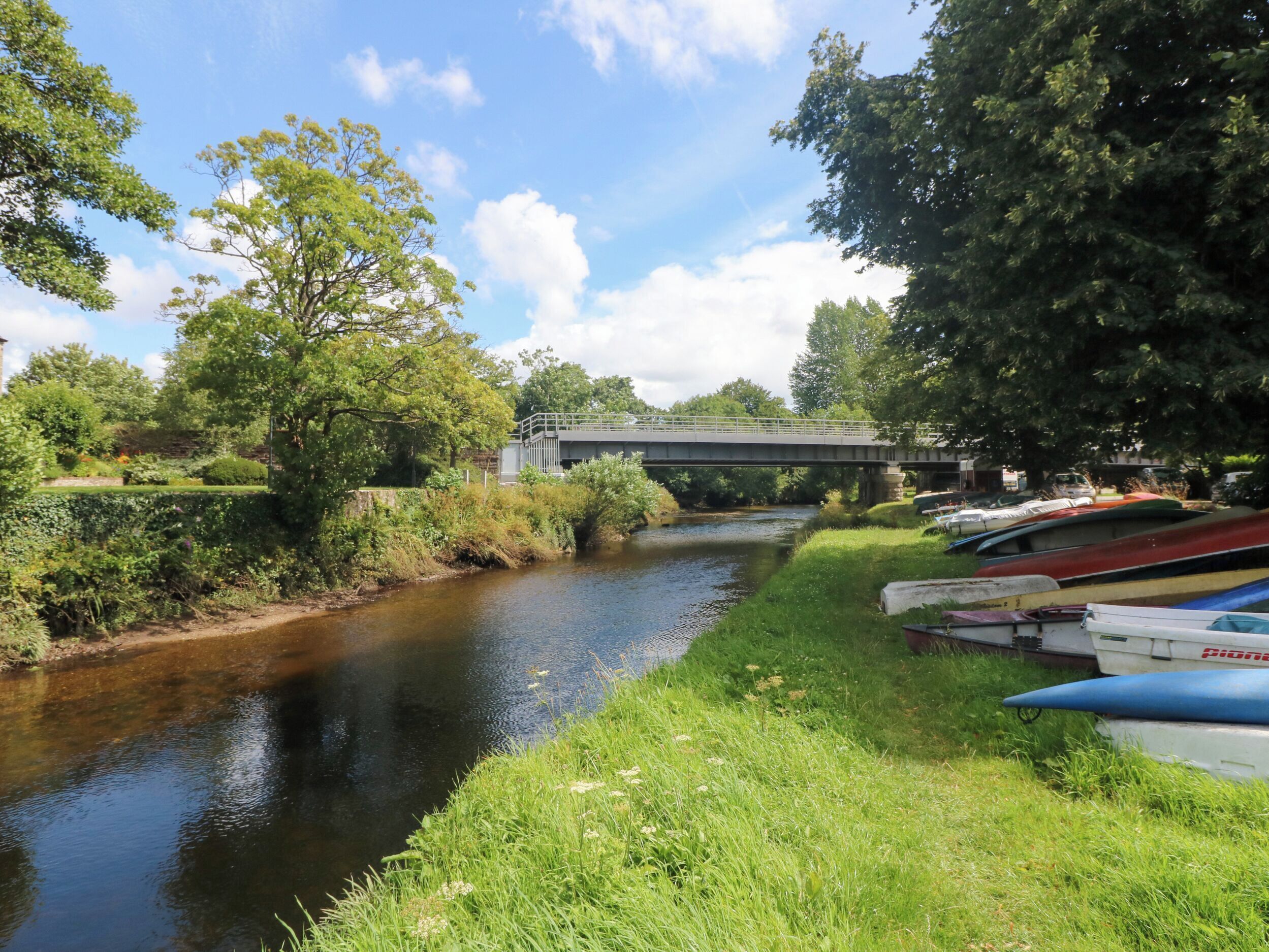 A river with boats docked on the right, a bridge in the background, and lush greenery on both sides.