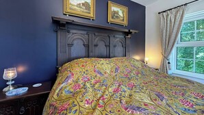 Een slaapkamer met een donker houten hoofdeinde, twee ingelijste foto’s aan de muur, een bed met een kleurrijk beddegoed en een nachtkastje met een lamp, plus een raam met gordijnen.