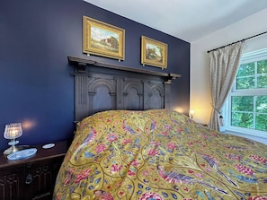 Un dormitorio con cabecera de madera oscura, dos cuadros en la pared, una cama con colcha de diseño colorido, una mesita de noche con lámpara y una ventana con cortinas.