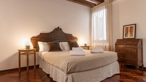 Un dormitorio con cabecera de madera, cama con ropa blanca, dos mesitas de noche con lámparas, cómoda de madera y ventana con cortinas translúcidas.