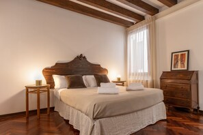 Ein Schlafzimmer mit einem Holzkopfteil, einem Bett mit weißen Bettwäsche, zwei Nachttischen mit Lampen, einer Holzkommode und einem Fenster mit durchsichtigen Vorhängen.