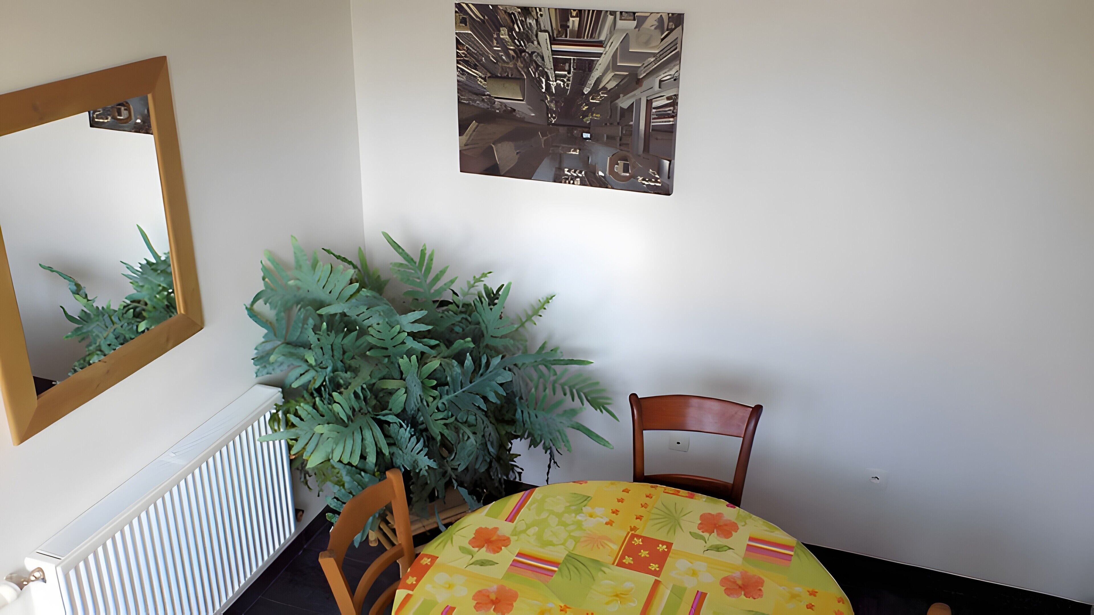 A room with a radiator, a mirror, a potted plant, a chair, a table with a floral tablecloth, and two wall-mounted pictures.