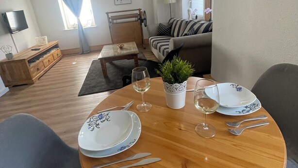 Uma sala de jantar com uma mesa de madeira redonda posta para dois, uma planta em vaso e um sofá ao fundo.