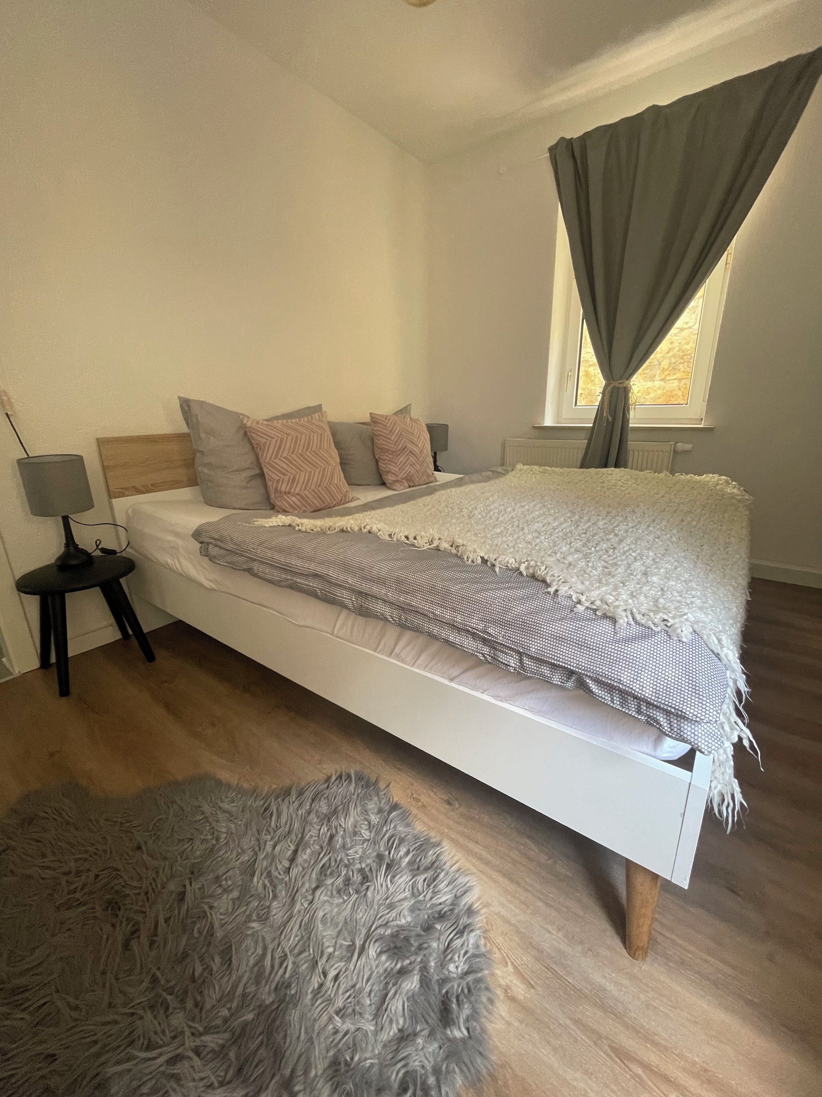 Ein Schlafzimmer mit Bett, Nachttisch, Lampe, einem Fenster mit Vorhängen und einem flauschigen Teppich.