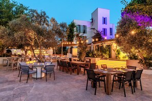 Un espacio exterior para comer con varias mesas y sillas, rodeado de árboles y un edificio de diseño moderno.