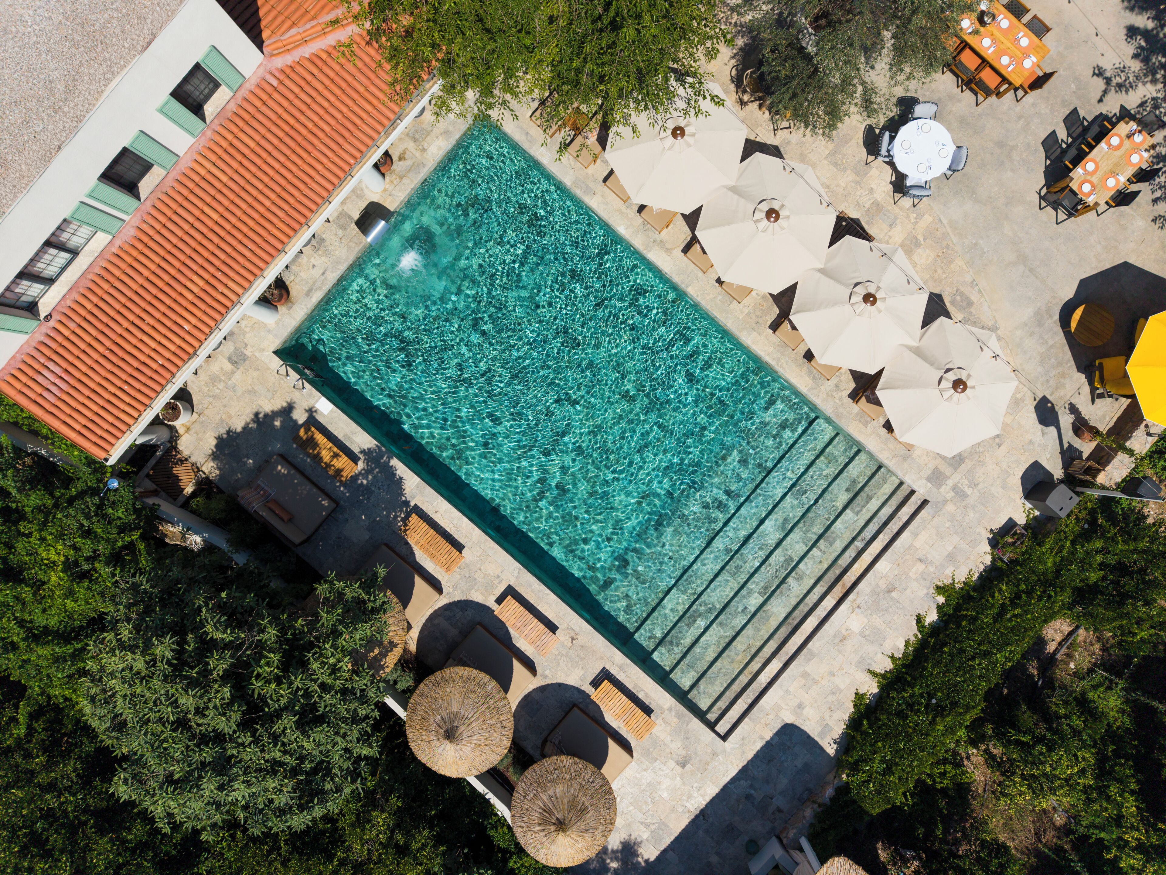 An aerial view of a swimming pool with surrounding lounge chairs and umbrellas, surrounded by trees and buildings.