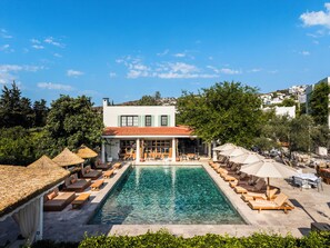 Un área junto a la piscina con sillones de descanso y sombrillas, un edificio con tejado de tejas rojas y un cielo azul despejado.
