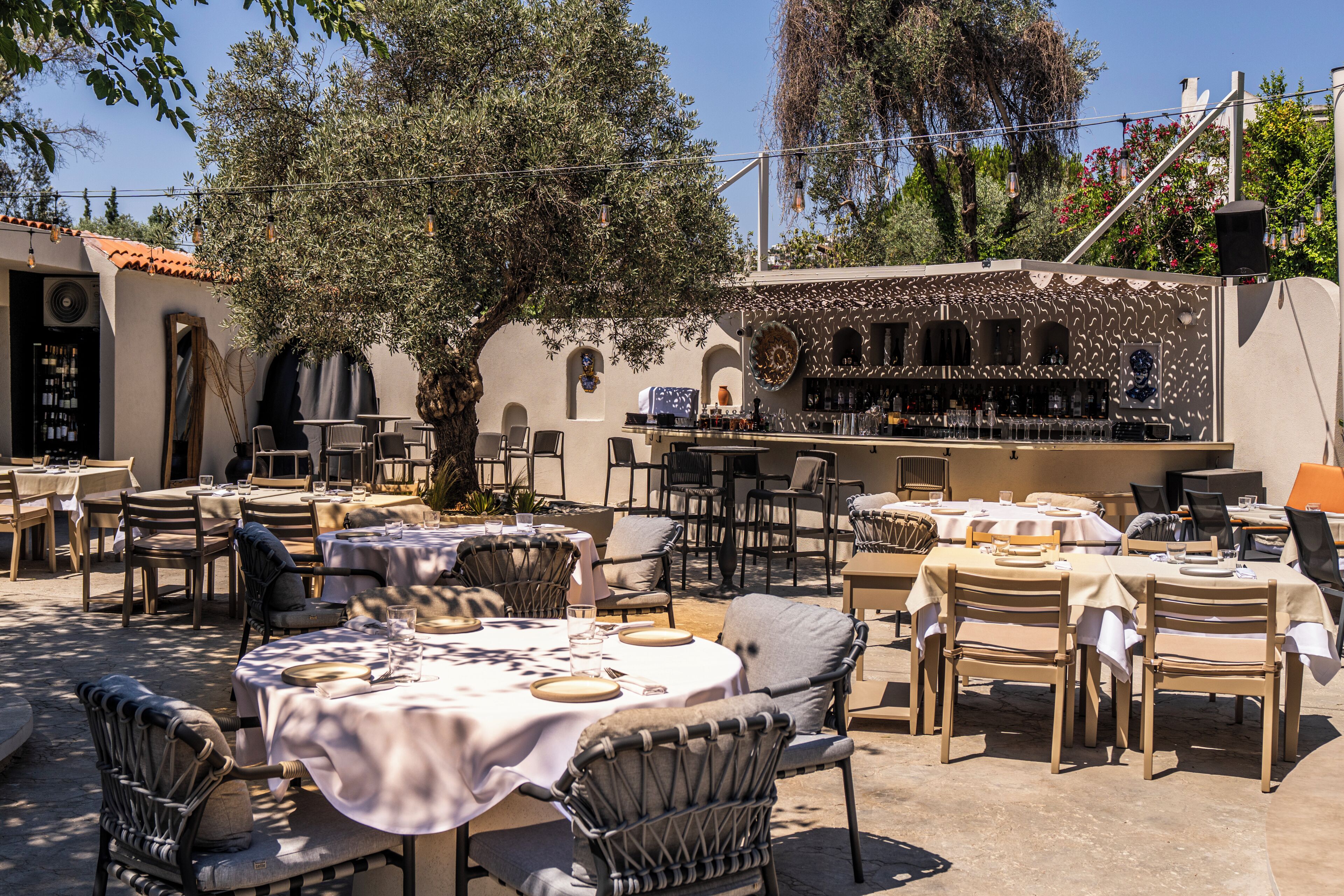 An outdoor dining area with tables and chairs, a bar, and a view of trees and buildings.
