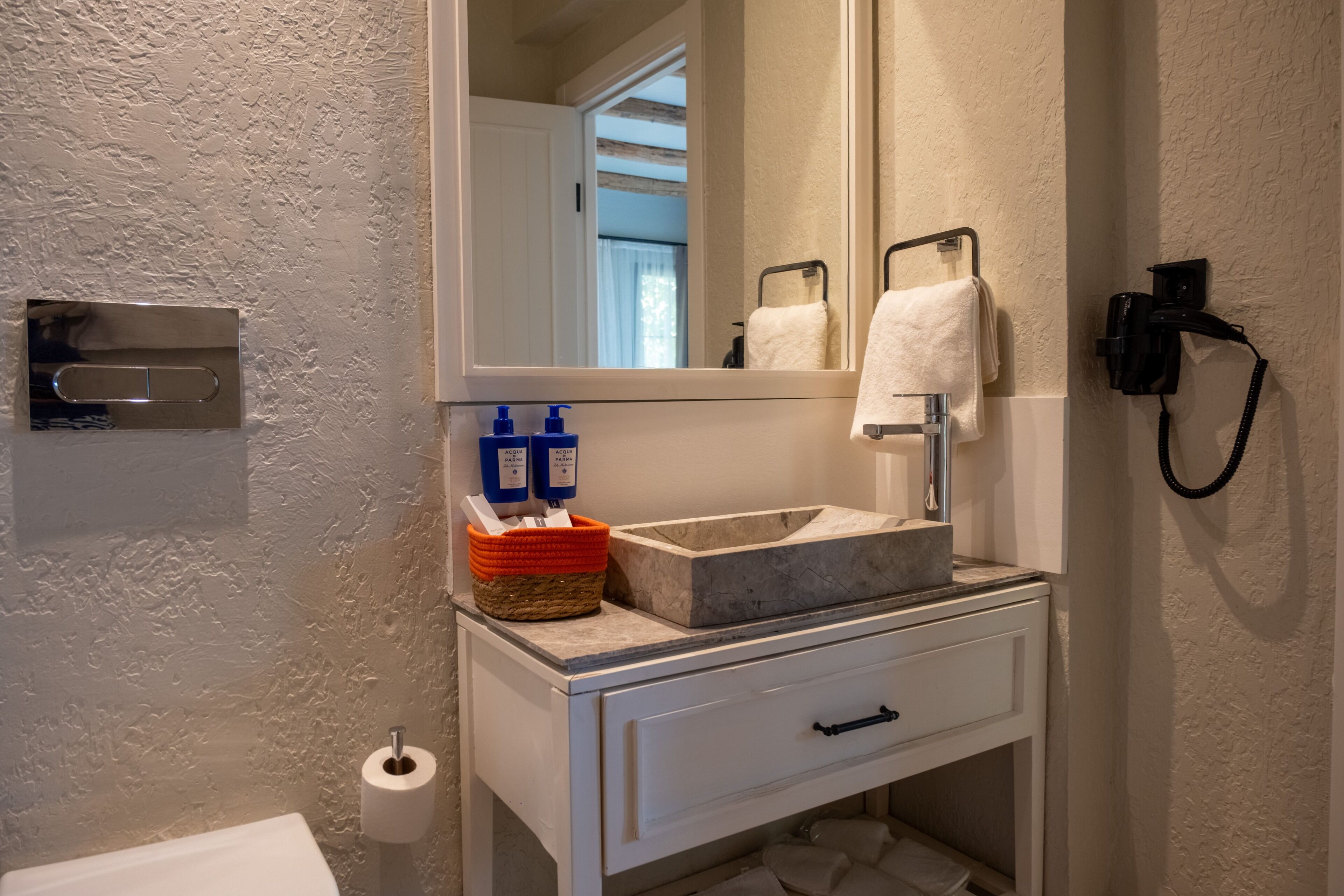 Un baño con lavamanos de mármol, un mueble blanco con cajón, toallero, secador de manos y porta papel higiénico.