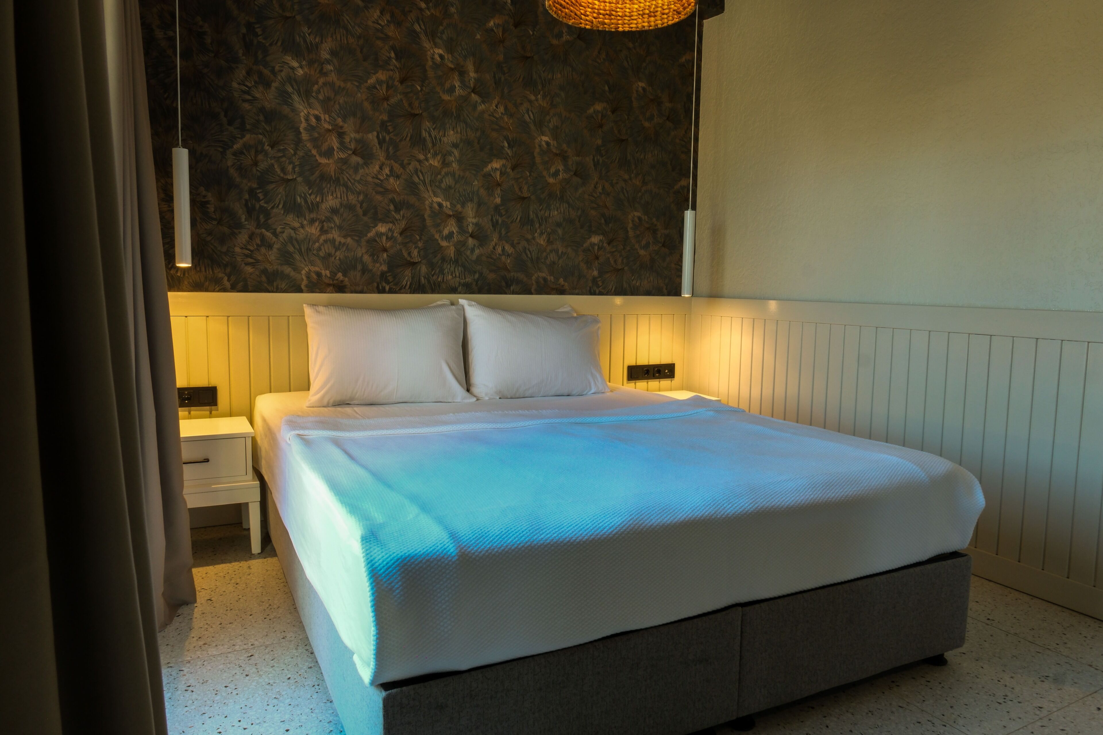 Een net opgemaakt bed met een wit hoofdeinde en kussens, een nachtkastje met een lamp, en een structuurwand achter het bed.