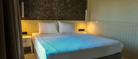 A neatly made bed with a white headboard and pillows, a bedside table with a lamp, and a textured wall behind the bed.