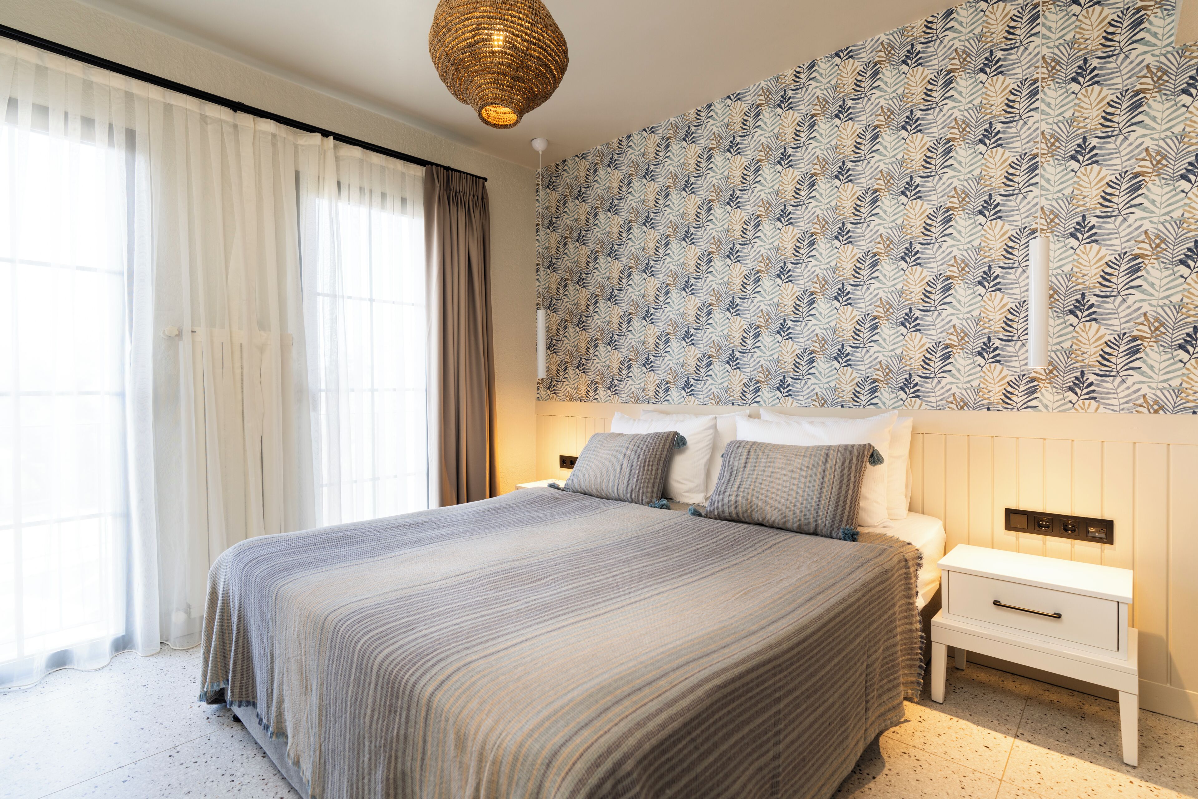 A bedroom with a large bed, a nightstand, a wall-mounted light fixture, and a patterned wallpaper.